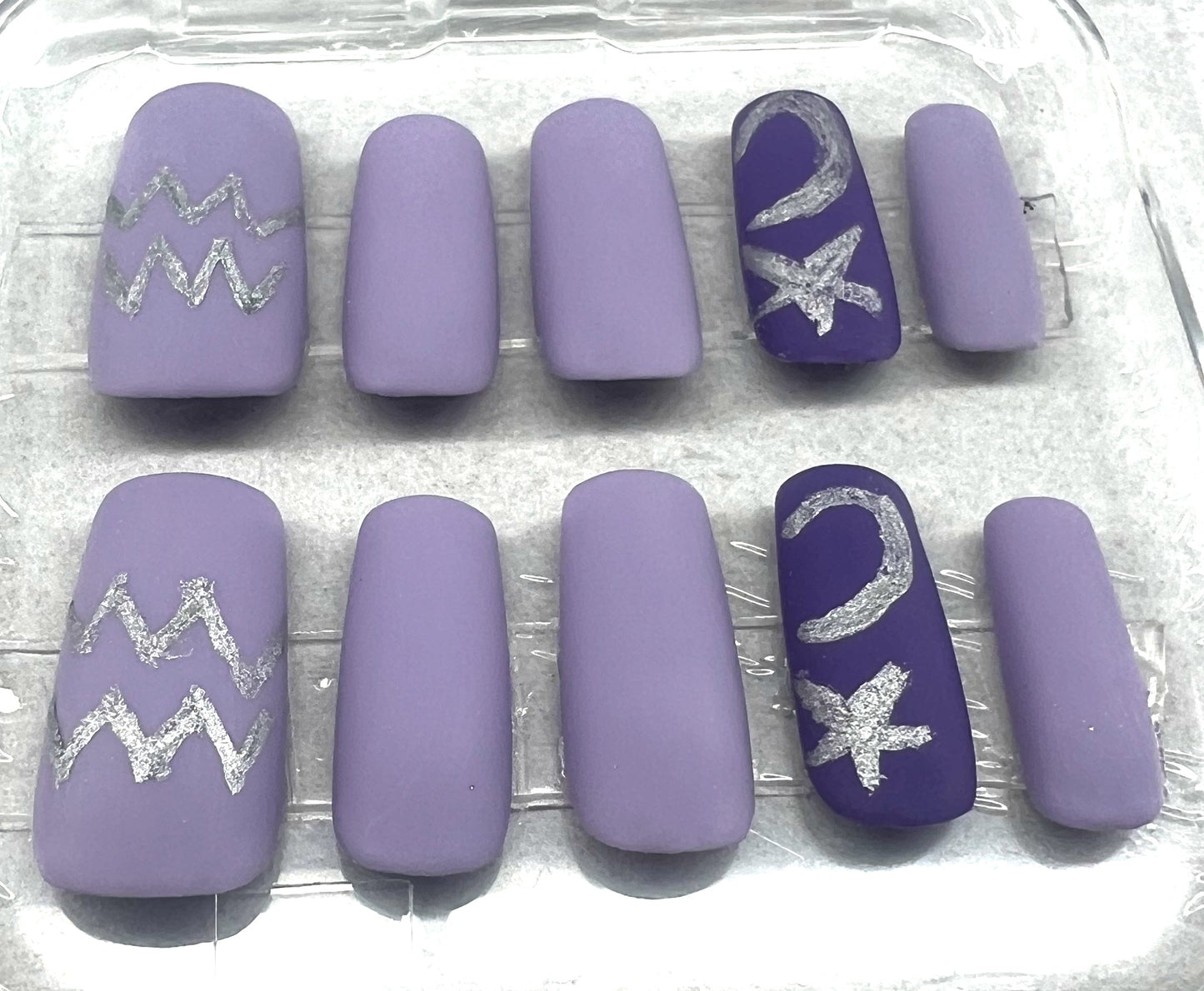 Aquarius at Dusk press-on nails in a soft-touch matte finish. The set features lavender nails with a dark purple accent nail on the ring finger. Hand-painted silver metallic zodiac symbols, stars, and moons are featured on the accent and thumb nails. Medium square shape.