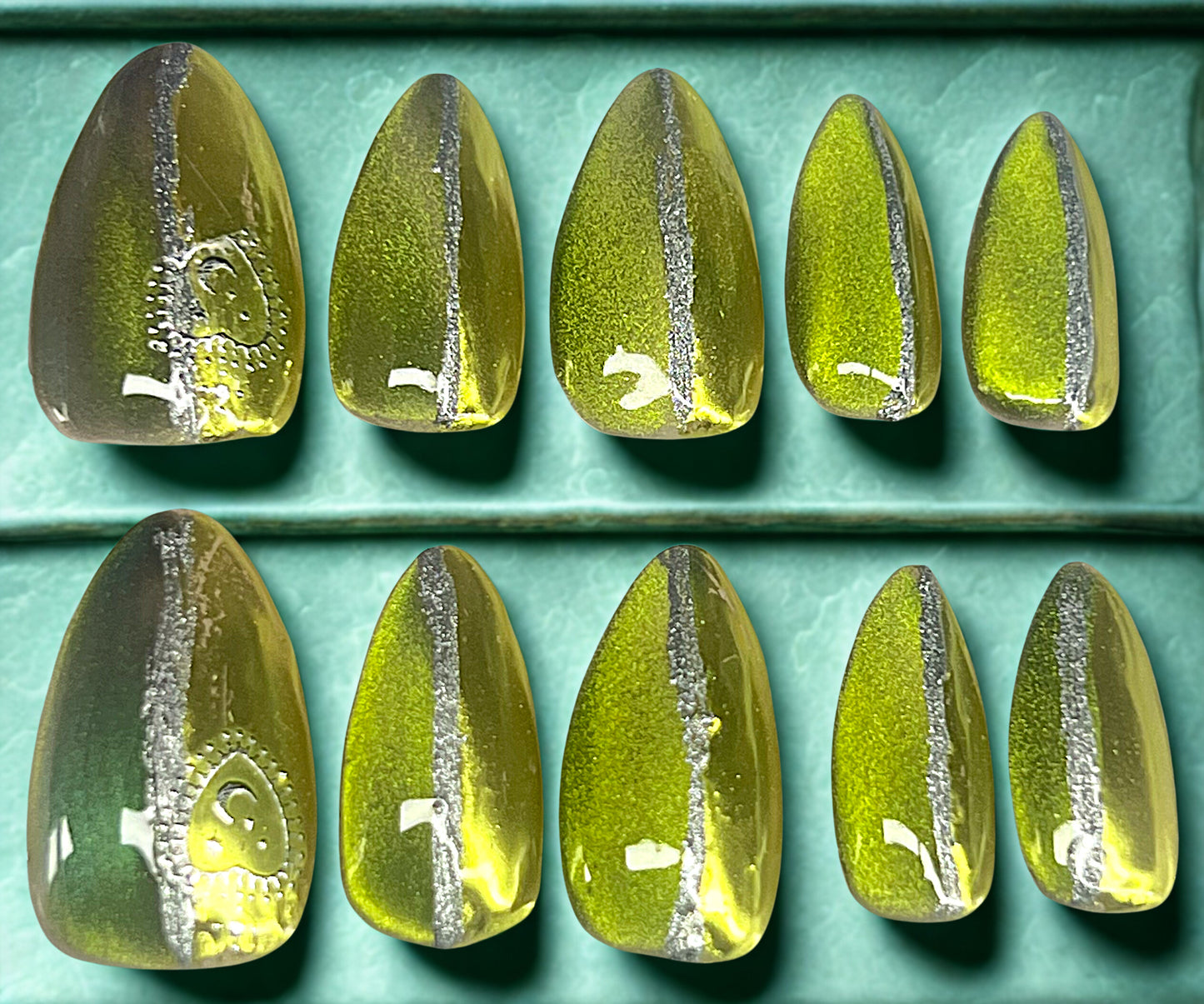 Astral Shift press-on nails featuring a black base with a split-finish design. One half is gold-green 9D velvet cat-eye and the other half is golden-green chrome, separated by a hand-painted vertical silver line. Silver celestial stickers on the thumbs. Medium almond shape.
