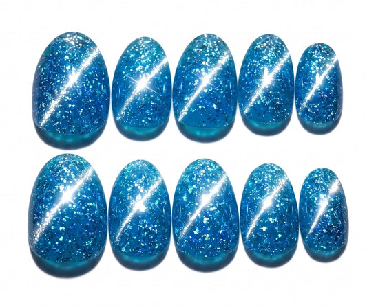 Cerulean blue jelly press on nails with a silver reflective glitter base that sparkles under light. The nails have a high-gloss finish and glow bright blue in the dark, displayed on a silver metal stand.