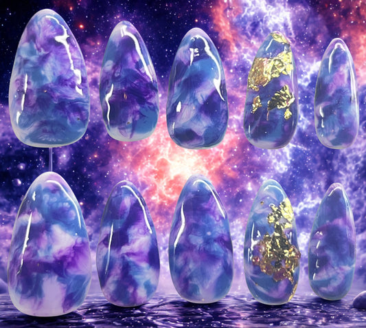 Cosmic Mist press-on nails featuring a white base with hand-painted dark blue and purple marble nebula effects. Ring fingers are decorated with textured gold foil. Medium almond shape shown against a vibrant purple and orange galaxy backdrop.