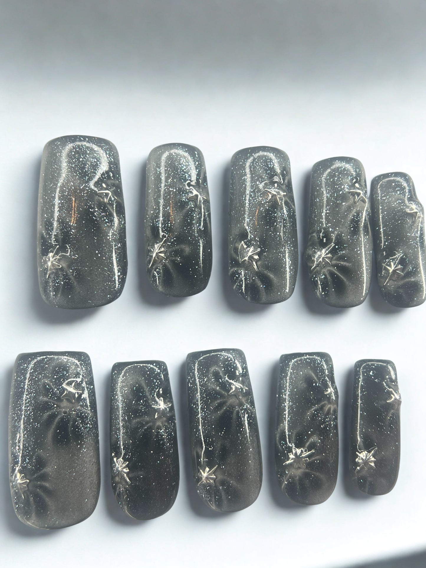  Five square press on nails with a black base featuring translucent glass cat's eye flowers and silver metallic star charms.