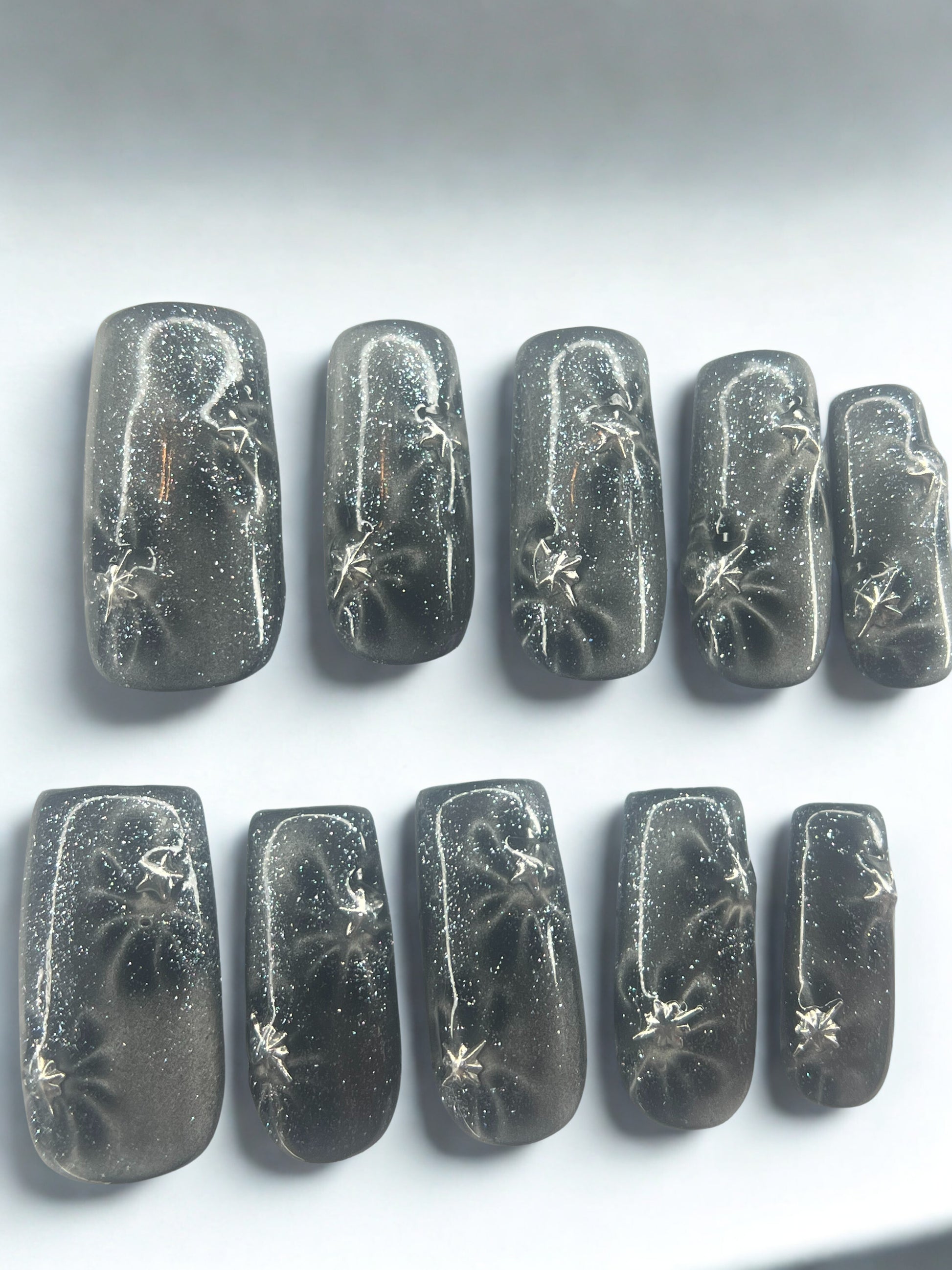  Five square press on nails with a black base featuring translucent glass cat's eye flowers and silver metallic star charms.