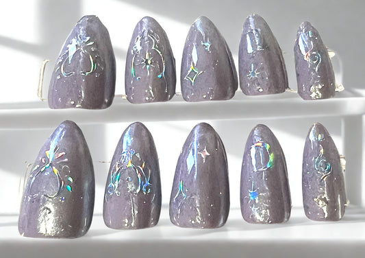 Medium length almond press-on nails in a soft lavender mermaid chrome finish. Each nail is decorated with prismatic holographic celestial stickers, including stars, crescents, and mystical symbols that reflect a rainbow of colors in the light.