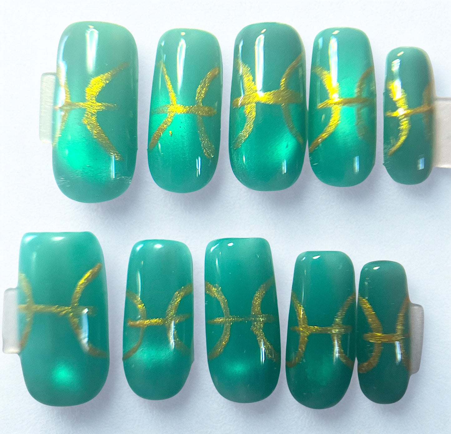 Teal jelly press-on nails with a magnetic cat-eye shimmer. Features hand-painted metallic gold Pisces zodiac symbols. The translucent milky white base gives a deep-sea glass effect on a square shape.