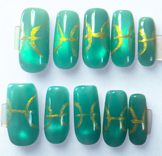 Teal jelly press-on nails with a magnetic cat-eye shimmer. Features hand-painted metallic gold Pisces zodiac symbols. The translucent milky white base gives a deep-sea glass effect on a square shape.