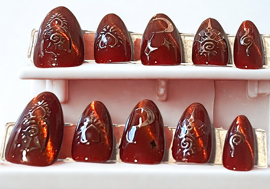 Handmade almond press-on nails in a rich 'liquid red' cat-eye finish. The magnetic shimmer creates a dimensional velvet look. Accented with structured rose gold metallic celestial symbols and moon charms. 