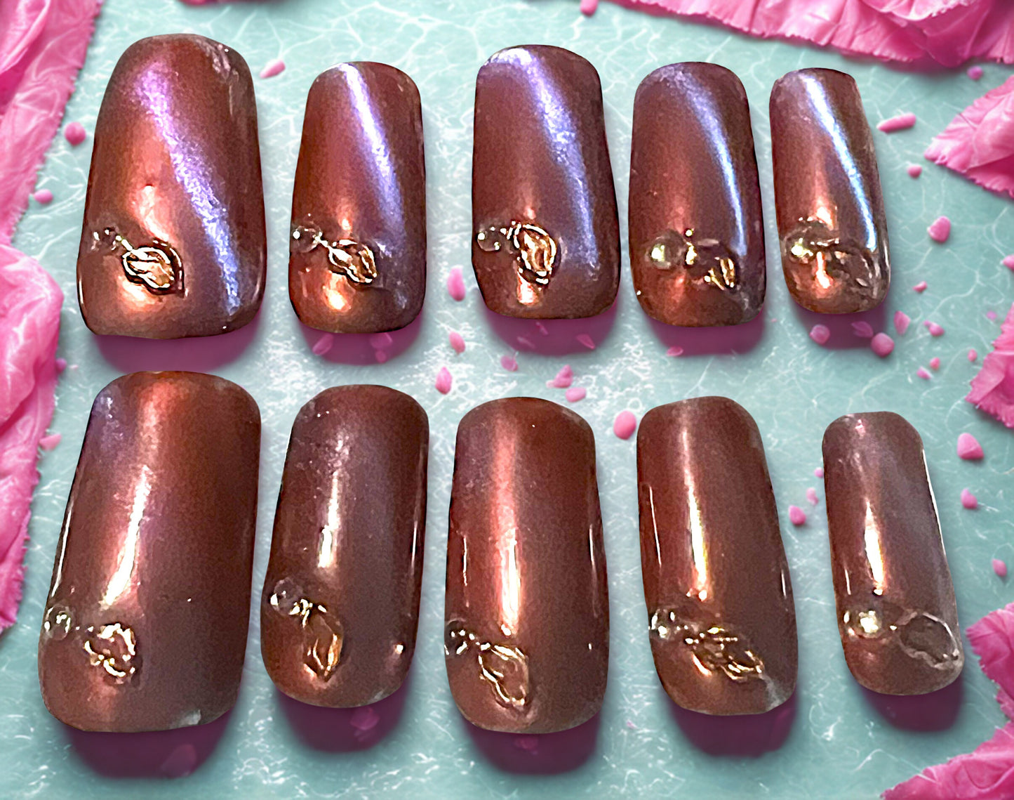 Golden Kiss press-on nails featuring a rose gold chrome finish over a lavender base. Includes a diagonal 9D cat-eye shimmer shifting between teal and pink. Each nail is decorated with a rose gold lip charm and one clear rhinestone. Medium square shape with a high-gloss metallic finish.