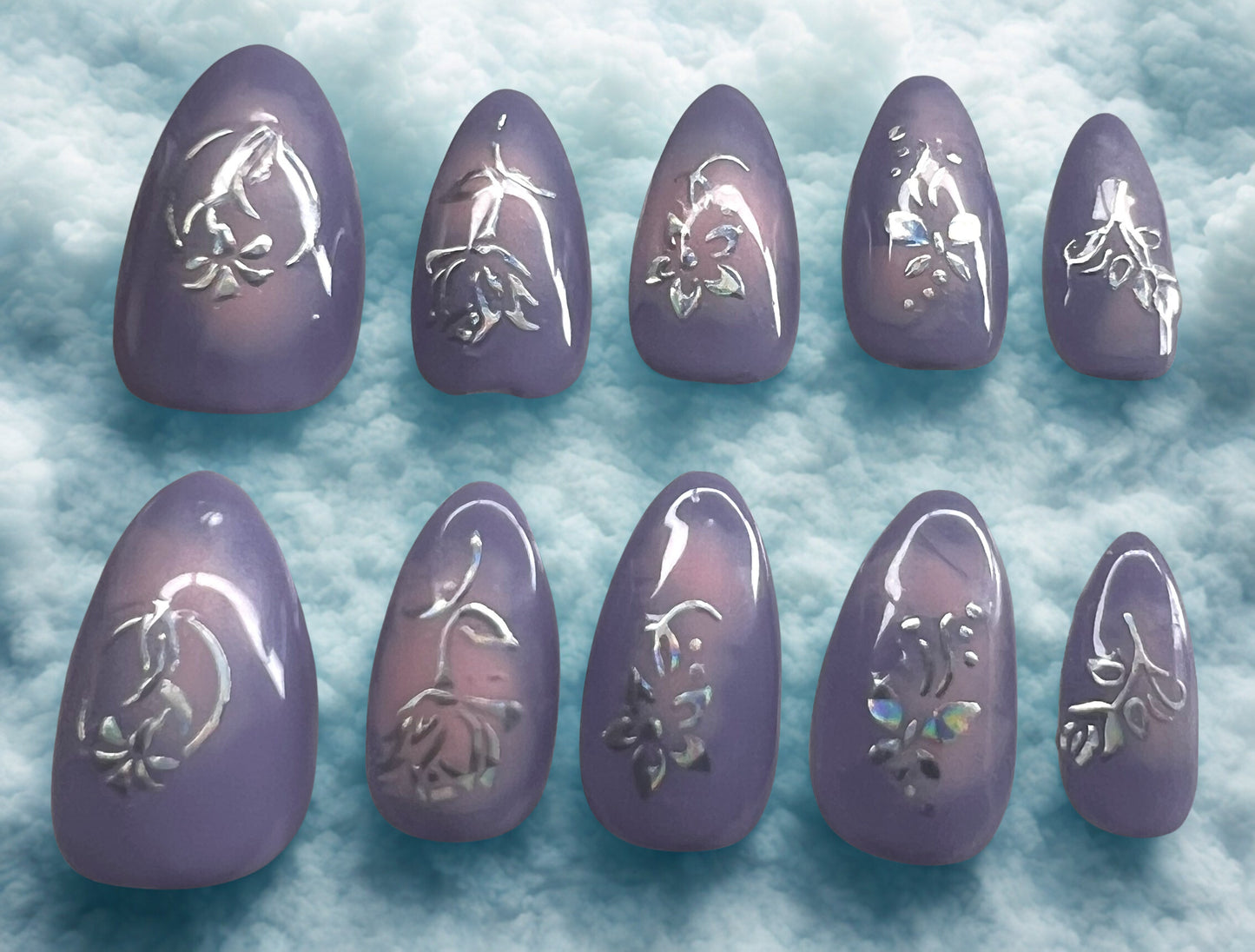 Blooming Love press-on nails featuring a lavender base with a soft pink aura bloomed in the center. Every nail is decorated with holographic flower and leaf stickers that reflect rainbow light. Short almond shape with a high-gloss finish against a cloud background.