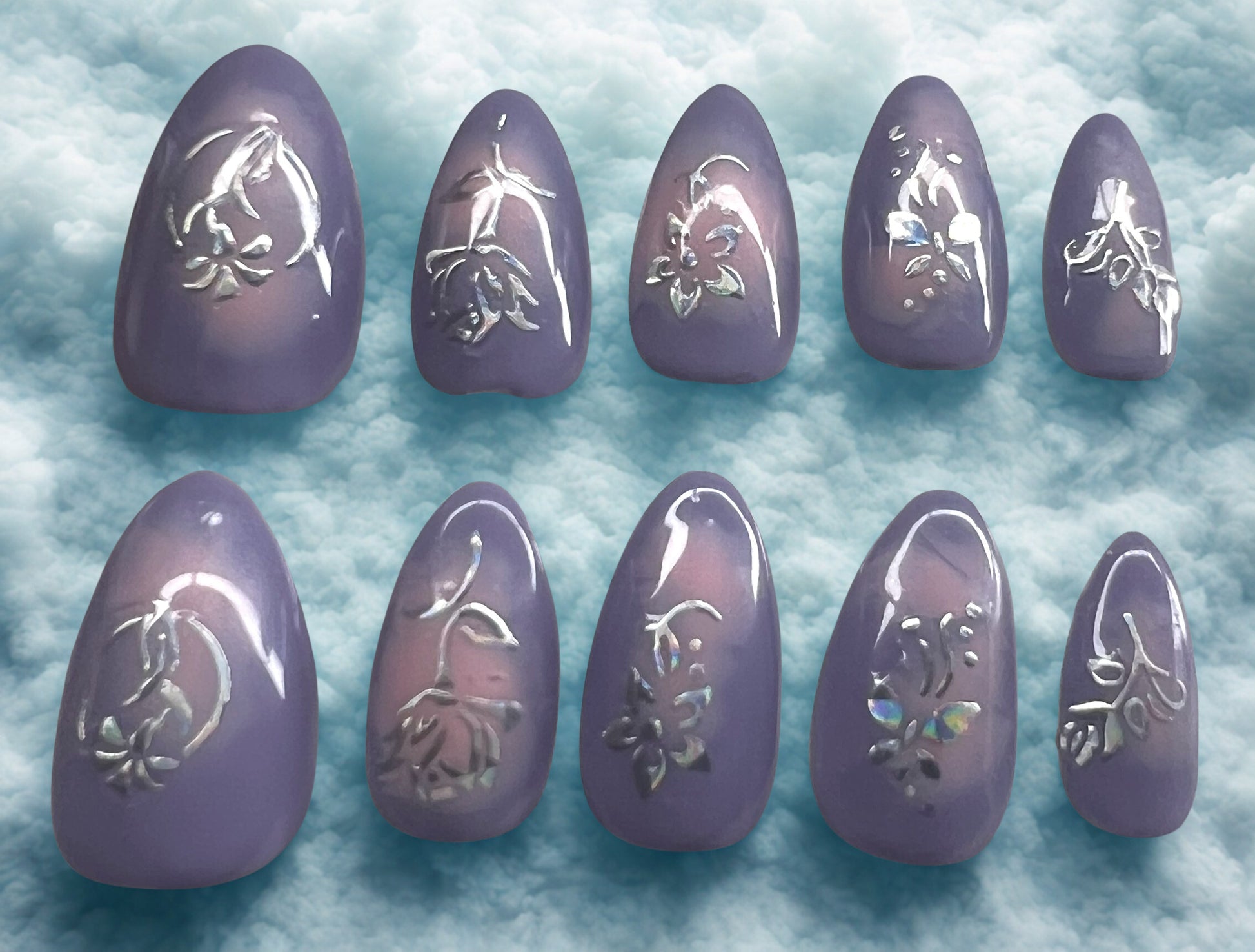 Blooming Love press-on nails featuring a lavender base with a soft pink aura bloomed in the center. Every nail is decorated with holographic flower and leaf stickers that reflect rainbow light. Short almond shape with a high-gloss finish against a cloud background.