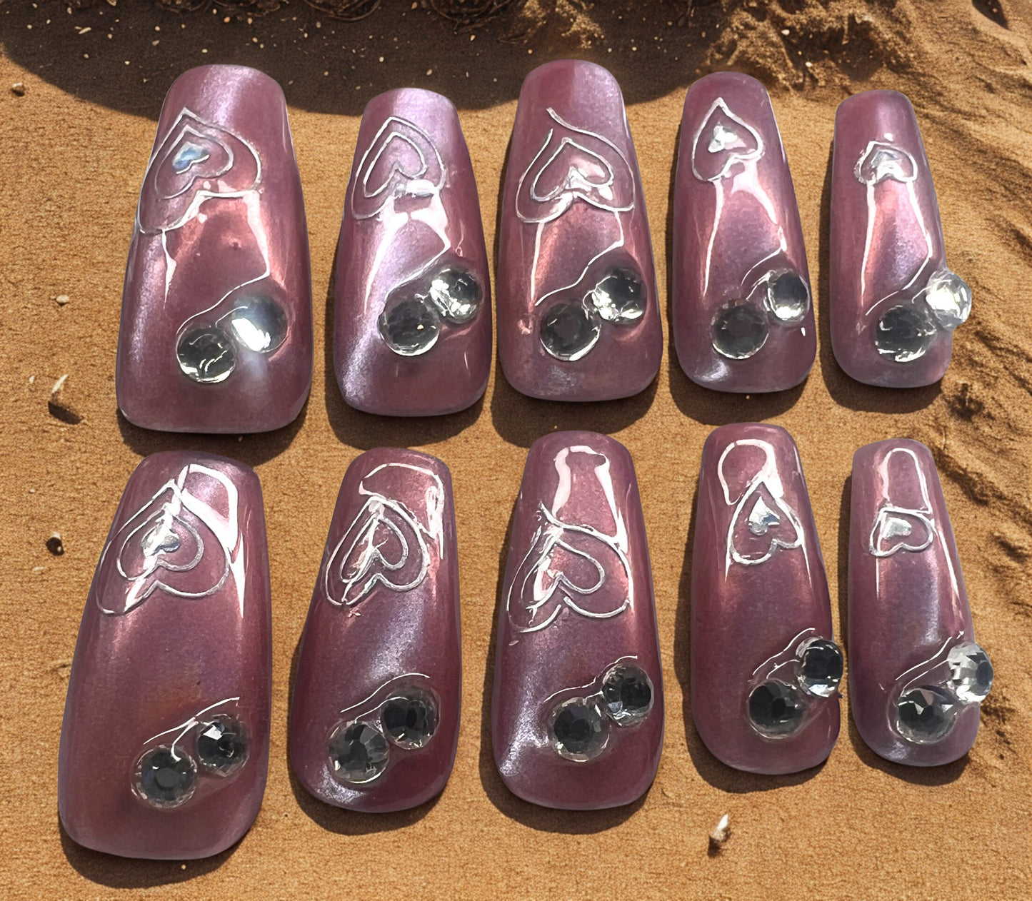 Rosewood Prism press-on nails featuring a nude base with rose gold chrome and sheer mermaid cat-eye shimmer. Decorated with holographic heart outlines and two clear rhinestones on each nail. Long coffin shape with a high-shine, prismatic finish.