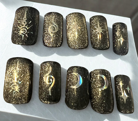 Obsidian Sky press-on nails featuring a black velvet cat-eye magnetic finish. Decorated with holographic celestial stickers including crescent moons, North stars, and sunburst symbols. Medium square shape with a high-shine finish.