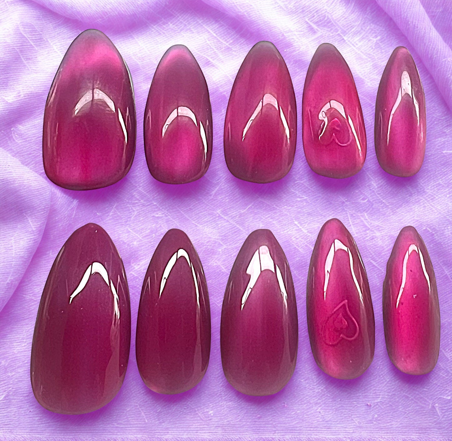 Hidden Love press-on nails featuring a lavender base with a glass velvet cat-eye shimmer. A holographic heart is encapsulated under a translucent pink jelly layer on the ring fingers. Medium almond shape against a purple silk fabric background.