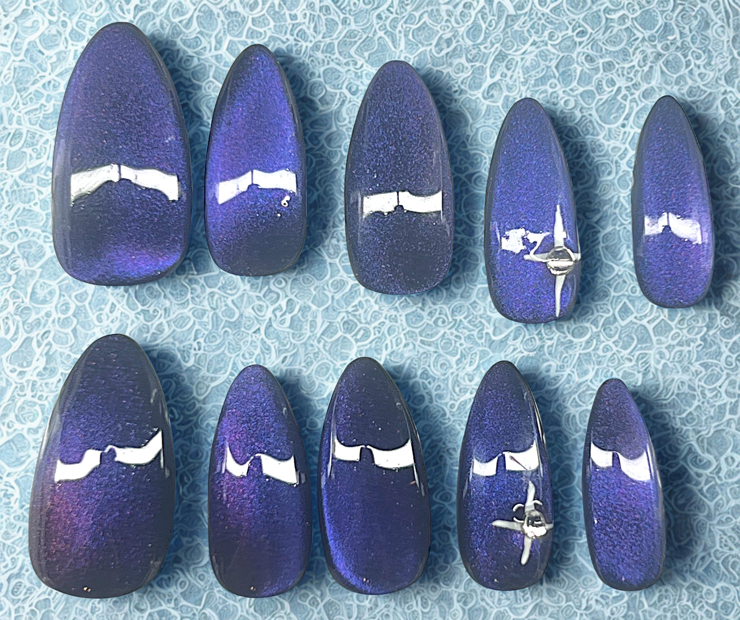 The Pulsar press-on nails featuring a black base with a 9D velvet cat-eye shifting through purple and blue. Ring fingers are decorated with a hand-painted white 4-point star and a single clear rhinestone. Medium stiletto shape against a textured light blue background.