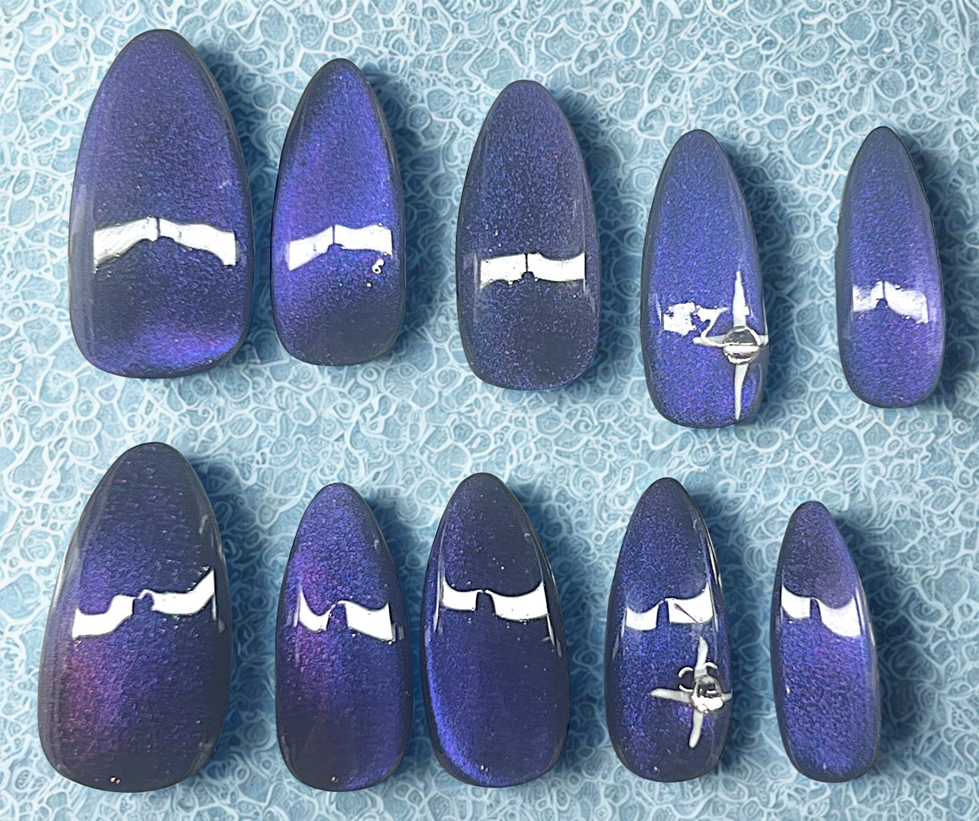 The Pulsar press-on nails featuring a black base with a 9D velvet cat-eye shifting through purple and blue. Ring fingers are decorated with a hand-painted white 4-point star and a single clear rhinestone. Medium stiletto shape against a textured light blue background.