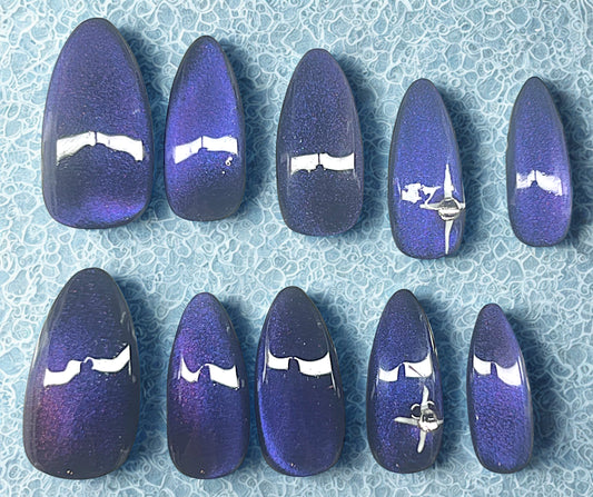 The Pulsar press-on nails featuring a black base with a 9D velvet cat-eye shifting through purple and blue. Ring fingers are decorated with a hand-painted white 4-point star and a single clear rhinestone. Medium stiletto shape against a textured light blue background.