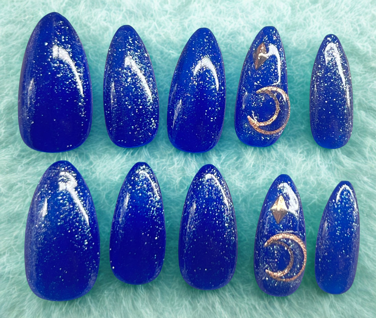 The Zenith press-on nails featuring a bright royal blue base with a fine silver glitter topcoat. Ring fingers are decorated with rose gold crescent moon and diamond-shaped charms. Medium almond shape shown against a fuzzy teal-blue background.
