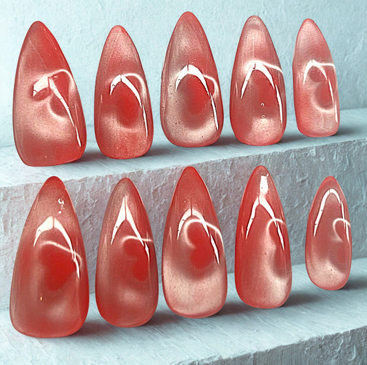 Velvet Heartbeat press-on nails featuring a red base with a glowing 'glass' cat-eye heart on every nail. The magnetic pigment isolates a sharp heart shape that appears to float inside the highly reflective, translucent finish. Medium stiletto shape on a light blue stone background.