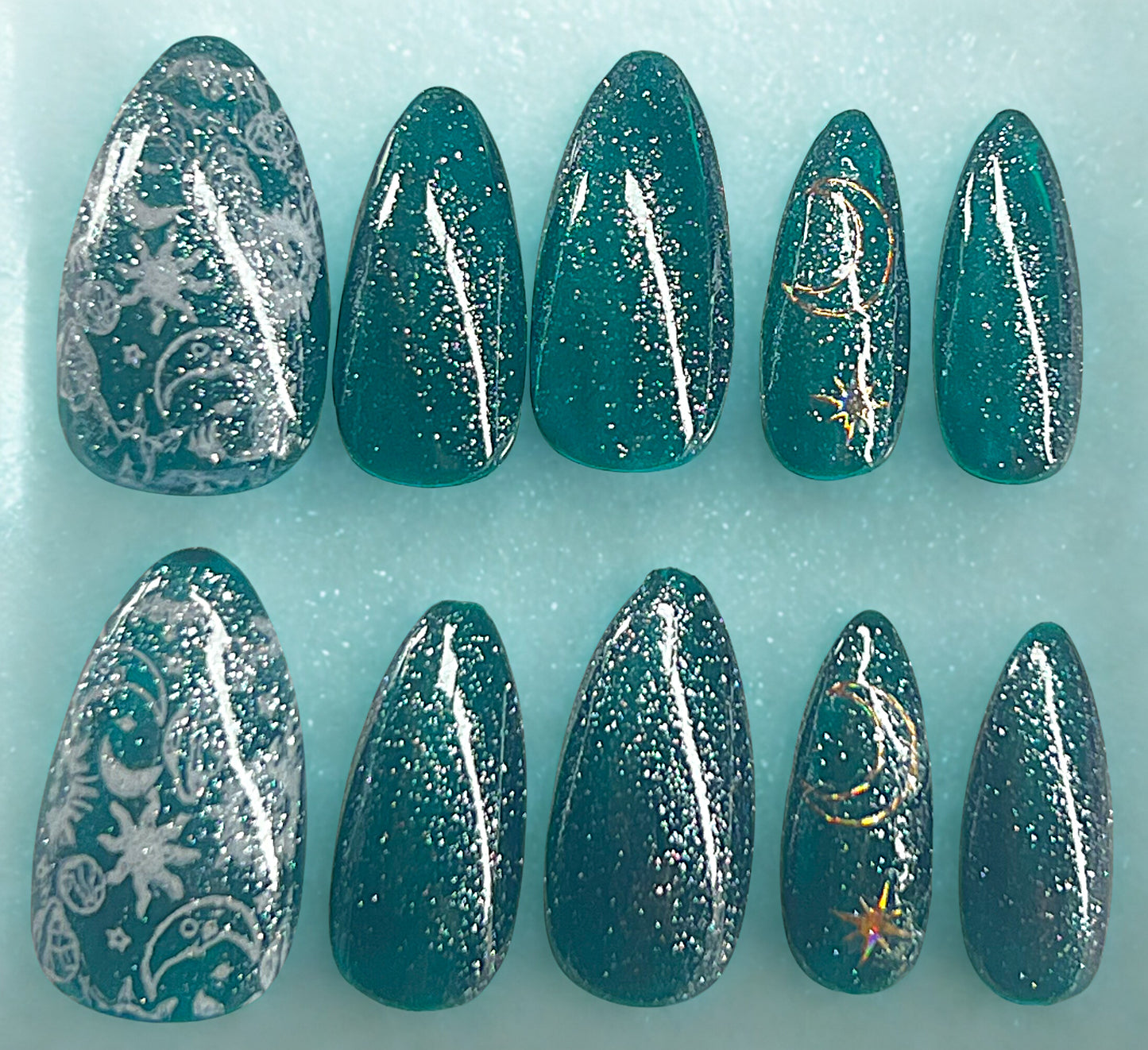 Deep Space press-on nails featuring a translucent green-turquoise jelly base with silver glitter topcoat. Thumbs feature white stamped celestial designs, and ring fingers include holographic gold-toned celestial stickers. Medium almond shape with a high-gloss, deep-sea finish.