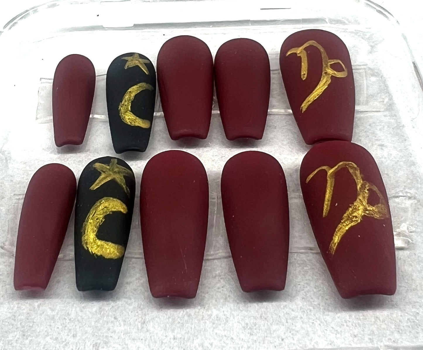 Capricorn at Midnight press-on nails in a velvet matte finish. The set features deep red nails with a matte black accent nail on the ring finger. Hand-painted silver metallic stars and zodiac symbols are featured on the black accent and red thumb nails. Medium coffin shape.