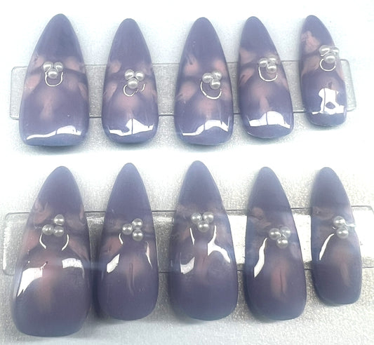 Handmade press on nails with a solid lavender purple base. Each nail features soft pink blooming flower art in the center, accented with three tiny white pearls. The nails are shown in a long pointed almond shape with a glossy topcoat.
