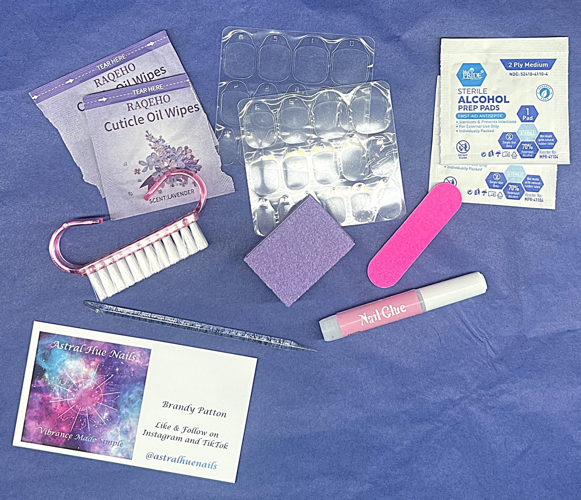 Astral Hue Deluxe Prep Kit spread out on a blue background. Includes a pink nail brush, purple buffer, pink file, adhesive tabs, nail glue, alcohol pads, cuticle oil wipes, a glittery cuticle stick, and a business card for Astral Hue Nails.