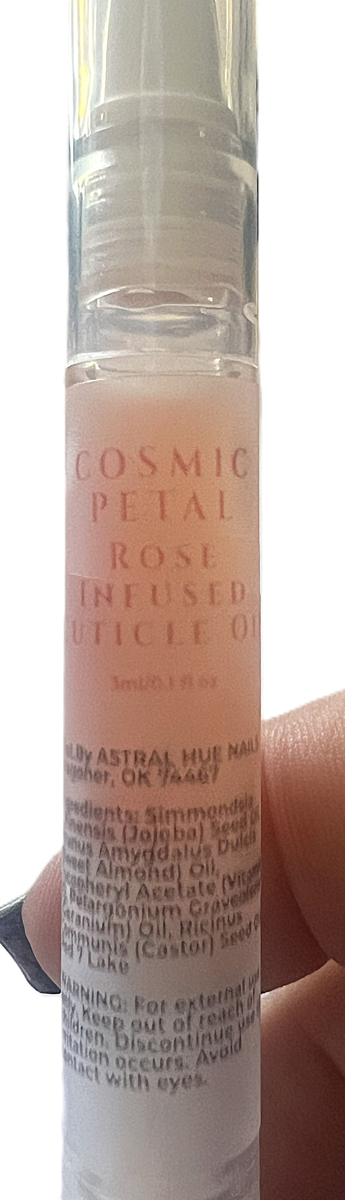 A close-up view of the 3ml Cosmic Petal Rose Infused Cuticle Oil twist pen by Astral Hue Nails, showing a clear bottle filled with a soft pink oil and a professional ingredient label listing jojoba oil, sweet almond oil, and castor oil.