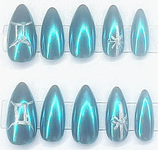 Ten handmade short almond press-on nails with a black base coated in an iridescent, metallic blue mirror chrome finish. The thumbs feature a hand-painted silver Gemini zodiac glyph, and the ring fingers feature a hand-painted silver celestial starburst, all presented on clear display stands against a clean background.