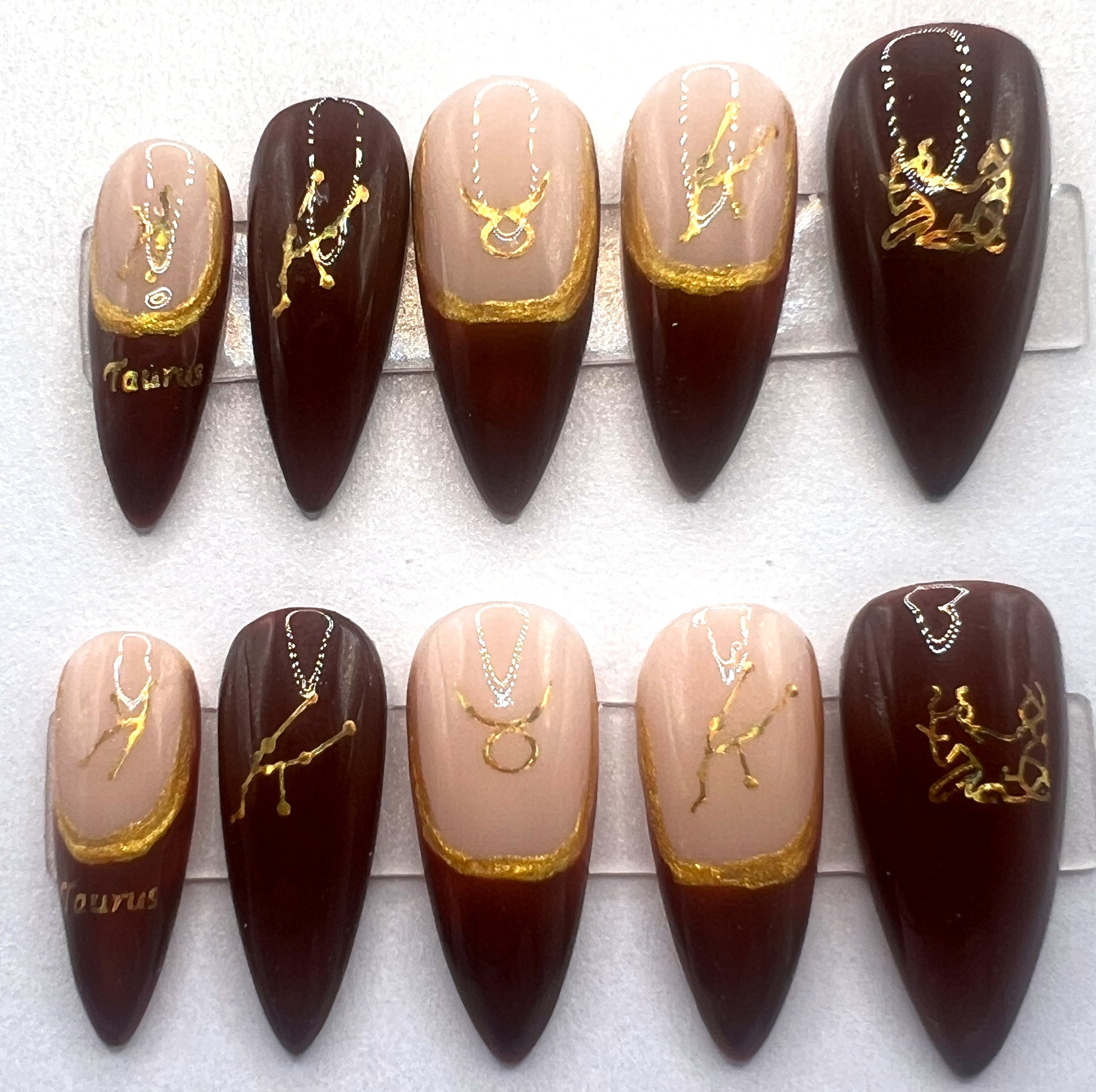 Hand-painted mismatched press on nails with a nude base and deep brown French tips. Features hand-painted gold outlines and gold holographic Taurus zodiac stickers on each nail. Shown in a medium stiletto shape.