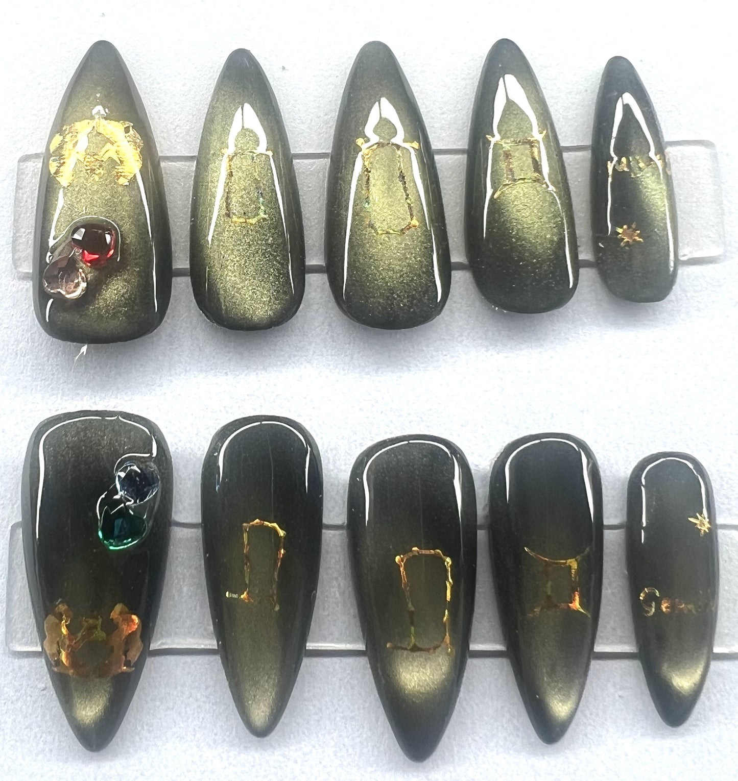 Ten handmade medium stiletto press-on nails featuring a black base with a shifting greenish-gold magnetic cat-eye shimmer. The thumbs are embellished with small, dimensional heart-shaped gemstones. All nails feature gold holographic celestial decals and fine star designs, displayed on clear stands against a clean background