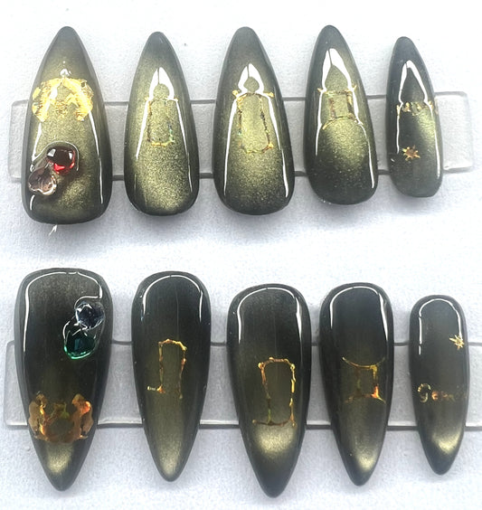 Ten handmade medium stiletto press-on nails featuring a black base with a shifting greenish-gold magnetic cat-eye shimmer. The thumbs are embellished with small, dimensional heart-shaped gemstones. All nails feature gold holographic celestial decals and fine star designs, displayed on clear stands against a clean background