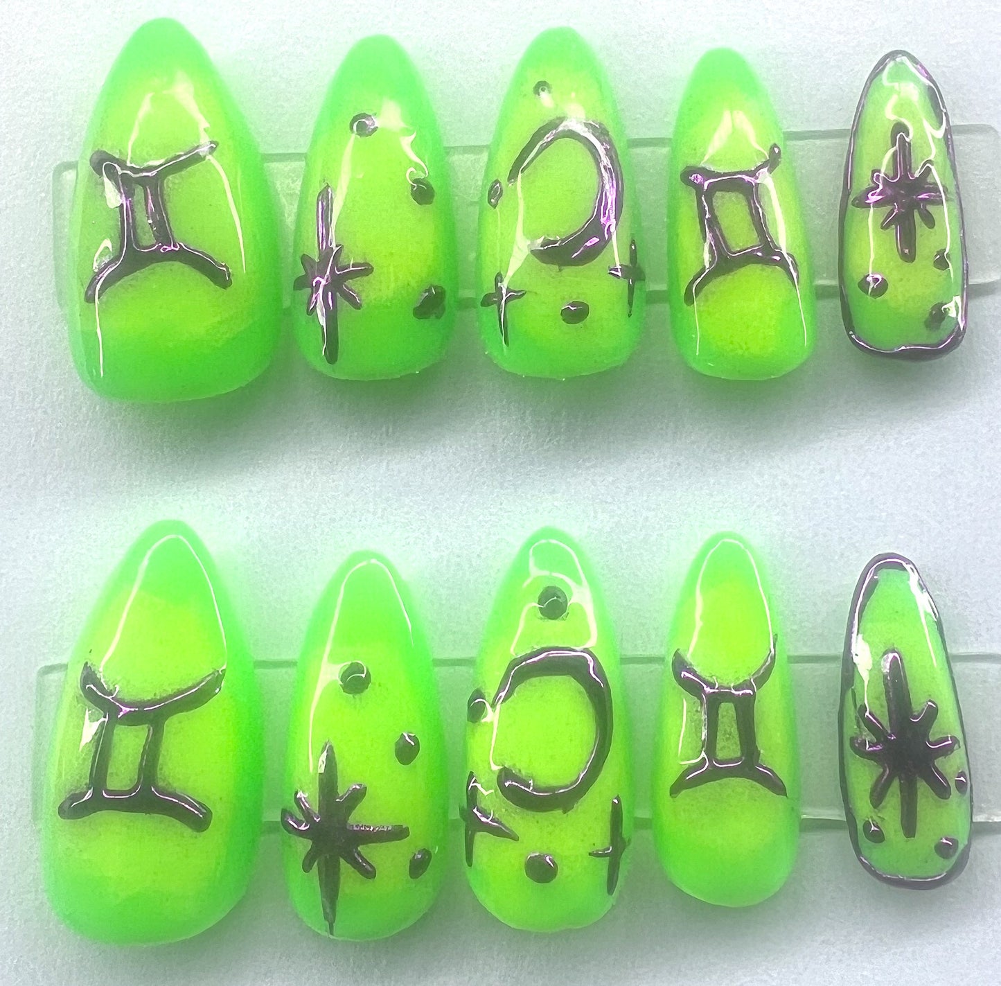 Ten handmade short coffin press-on nails featuring a neon green base color with a blended neon yellow aura gradient in the center. The surface is decorated with raised, hand-painted metallic purple chrome elements including the Gemini zodiac sign, multi-pointed stars, and crescent moons, shown on clear stands against a clean background.