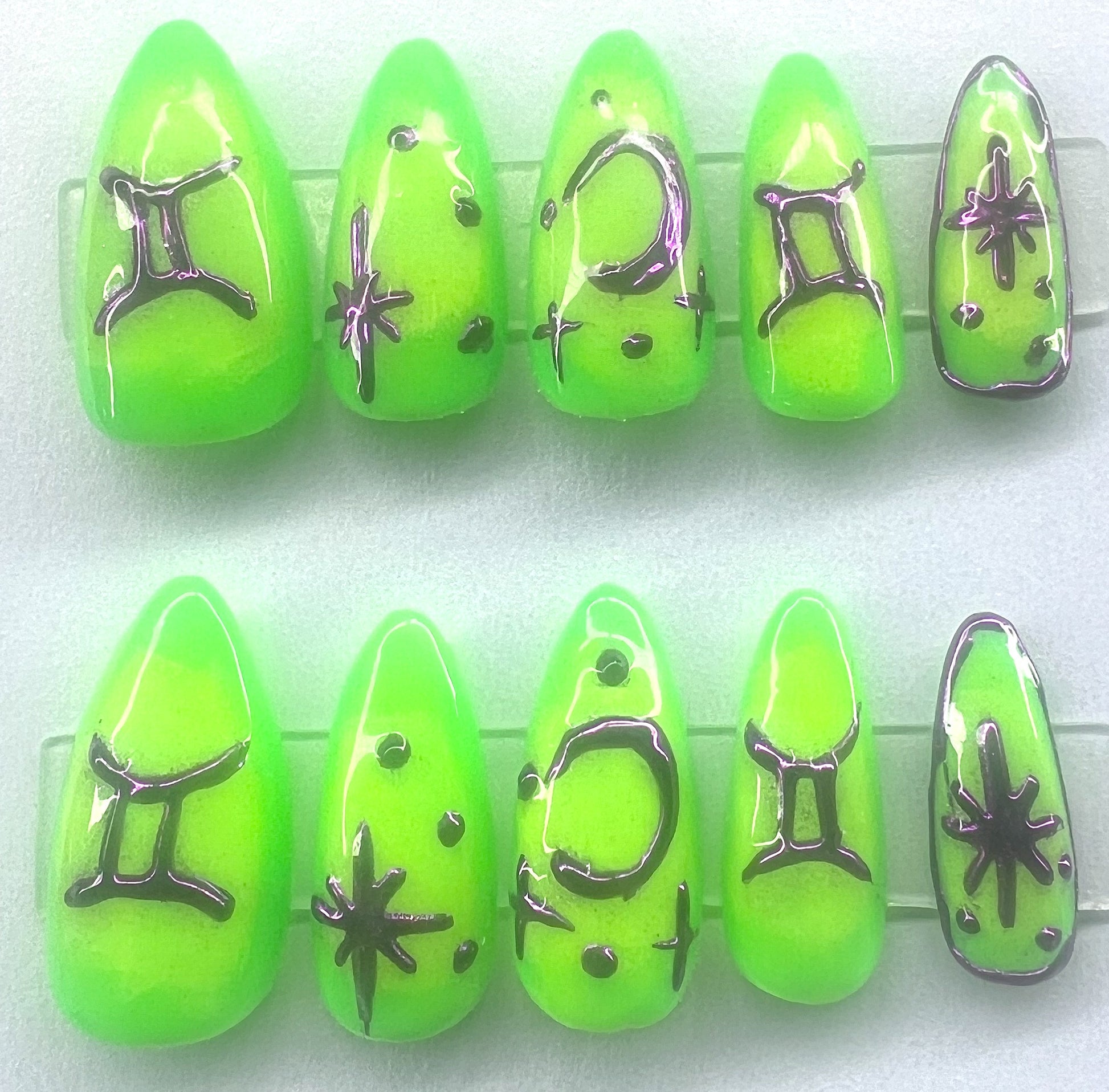 Ten handmade short coffin press-on nails featuring a neon green base color with a blended neon yellow aura gradient in the center. The surface is decorated with raised, hand-painted metallic purple chrome elements including the Gemini zodiac sign, multi-pointed stars, and crescent moons, shown on clear stands against a clean background.