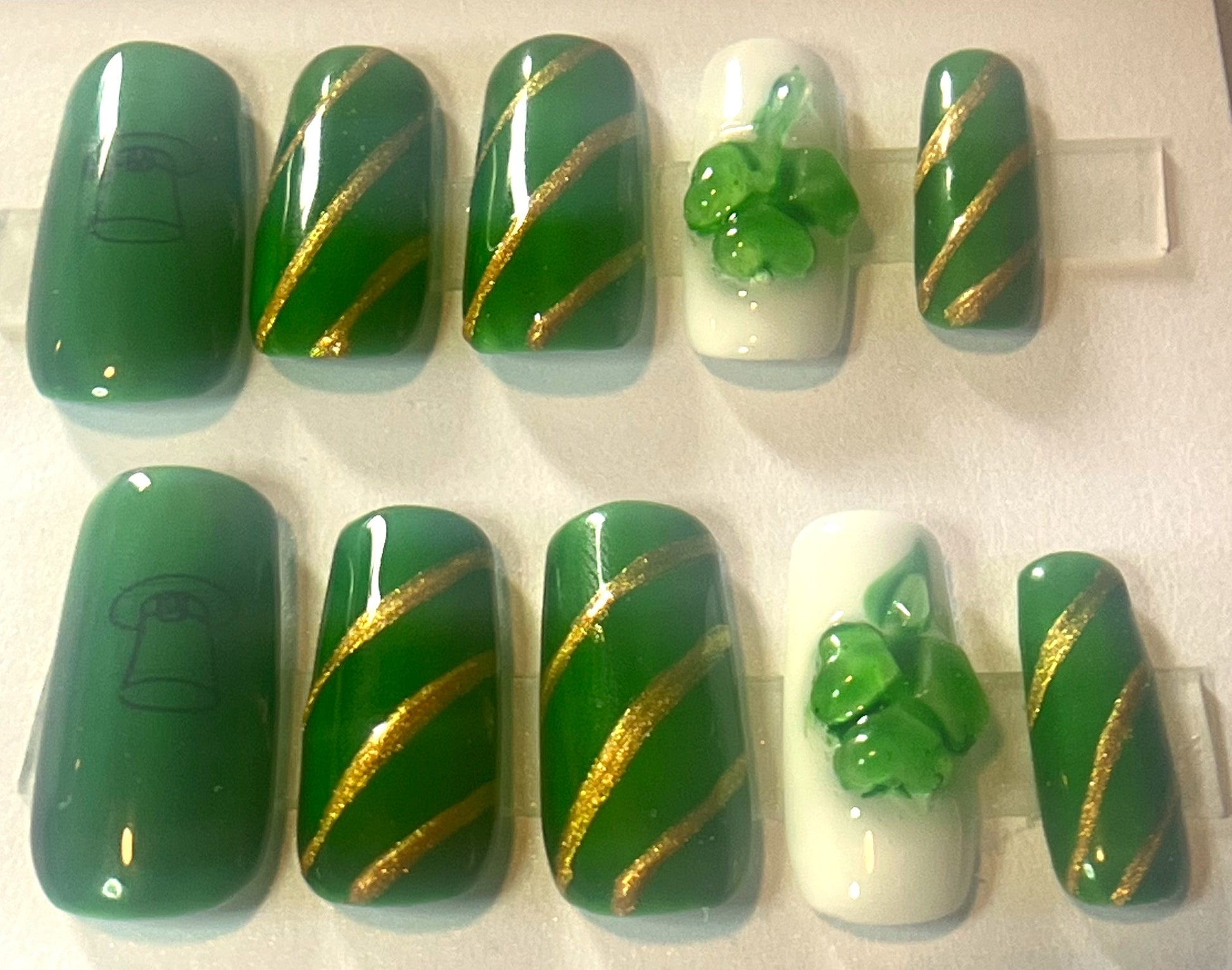 Close up of St. Patrick's Day press-on nails with a sheer emerald green jelly finish. Features 3D embossed green shamrocks and intricate gold metallic linework on a short coffin shape.