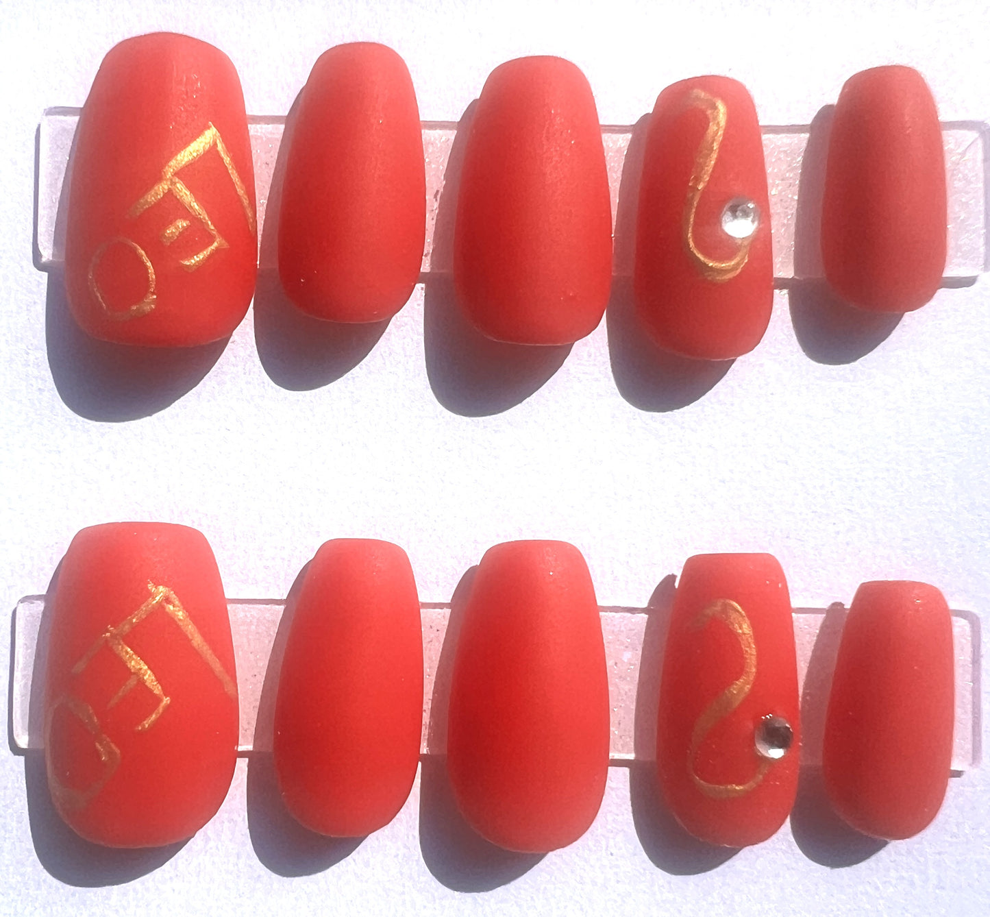 Short coffin-shaped press-on nails featuring a matte fire-red finish. The thumb nails display metallic gold linework spelling out LEO, while the ring fingers feature a hand-painted gold Leo zodiac symbol embellished with a small, clear sparkling rhinestone at the base.