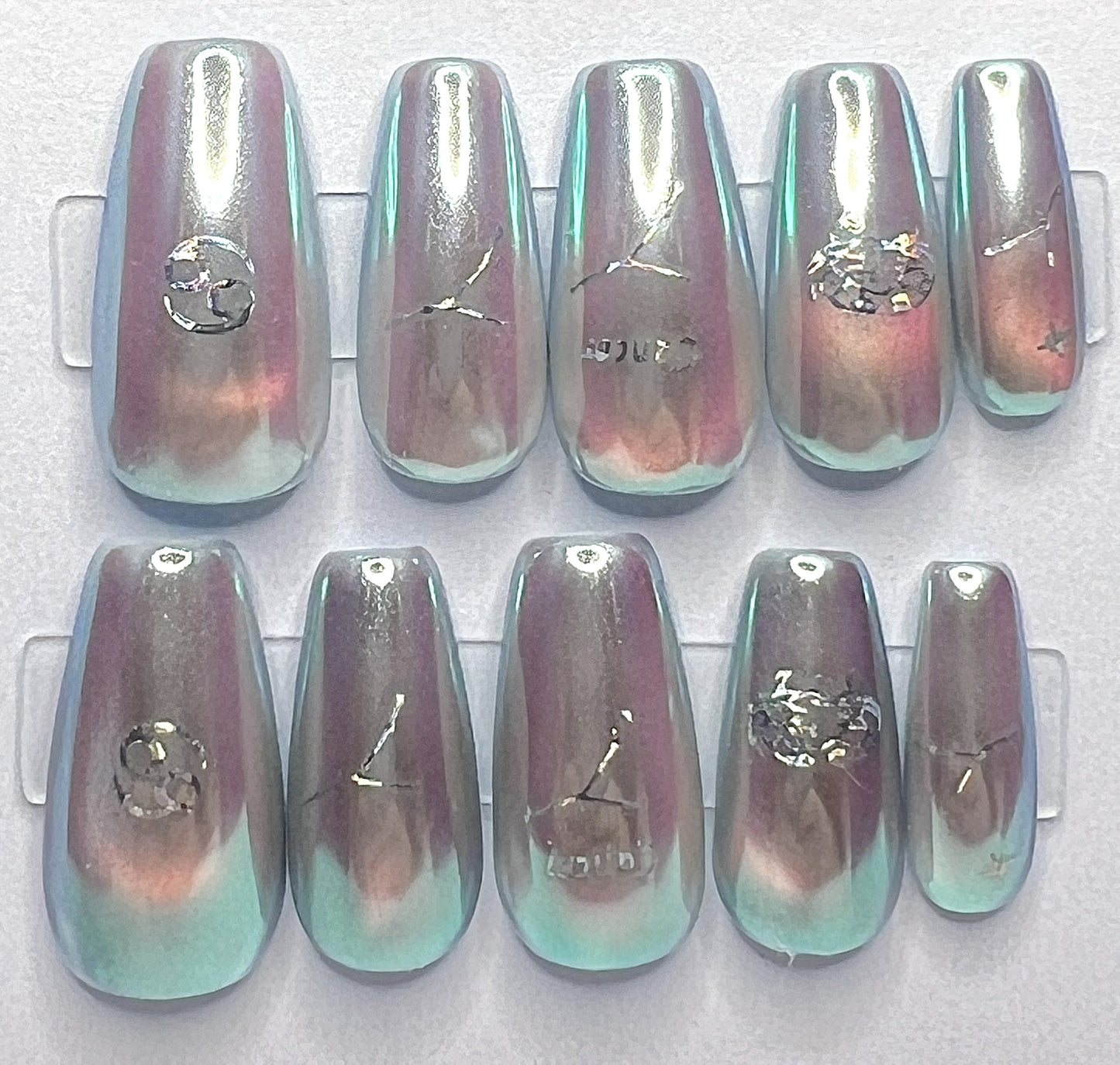 Medium coffin-shaped press-on nails featuring a pale blue base with a glowing glass cat-eye design underneath a high-shine, iridescent mermaid pearl chrome powder. Holographic silver Cancer zodiac symbols and constellation decals are placed cleanly on the centers of the metallic nails.