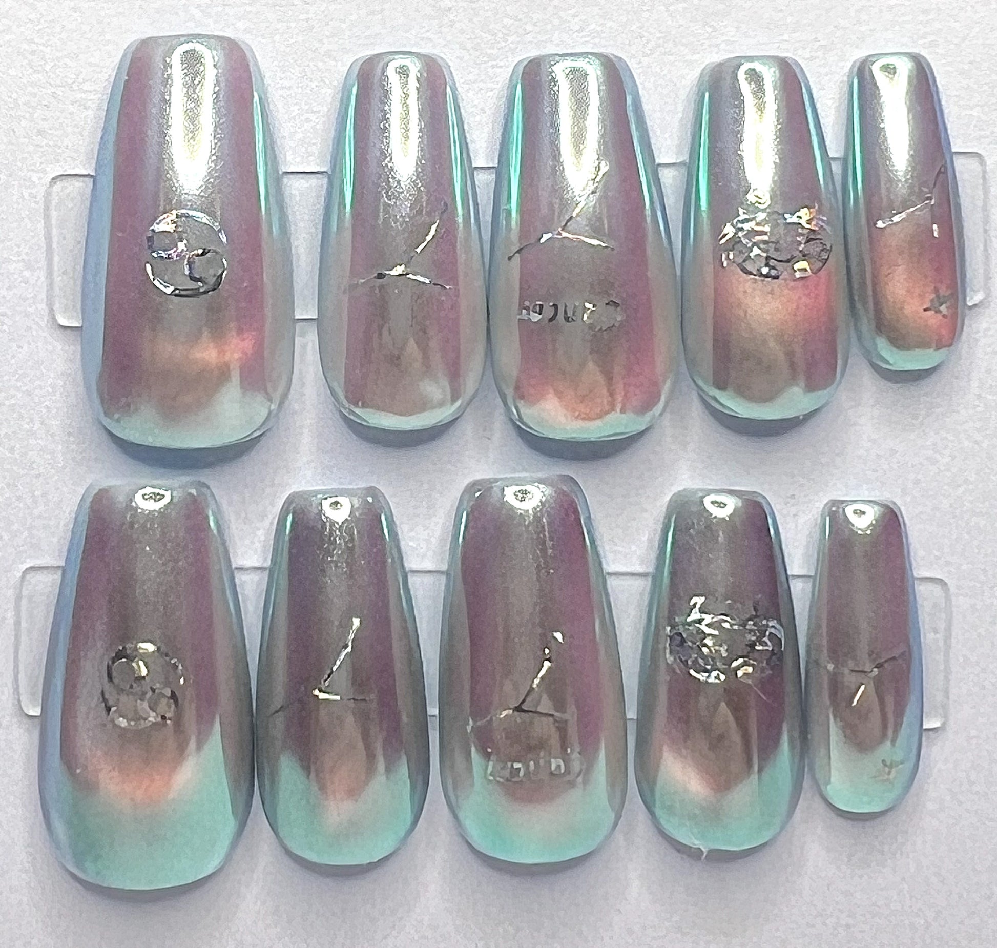 Medium coffin-shaped press-on nails featuring a pale blue base with a glowing glass cat-eye design underneath a high-shine, iridescent mermaid pearl chrome powder. Holographic silver Cancer zodiac symbols and constellation decals are placed cleanly on the centers of the metallic nails.