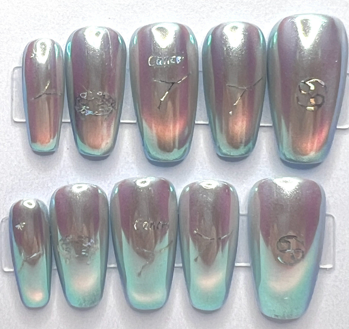 Medium coffin-shaped press-on nails featuring a pale blue base with a glowing glass cat-eye design underneath a high-shine, iridescent mermaid pearl chrome powder. Holographic silver Cancer zodiac symbols and constellation decals are placed cleanly on the centers of the metallic nails.