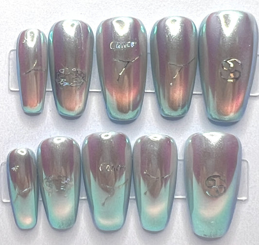 Medium coffin-shaped press-on nails featuring a pale blue base with a glowing glass cat-eye design underneath a high-shine, iridescent mermaid pearl chrome powder. Holographic silver Cancer zodiac symbols and constellation decals are placed cleanly on the centers of the metallic nails.