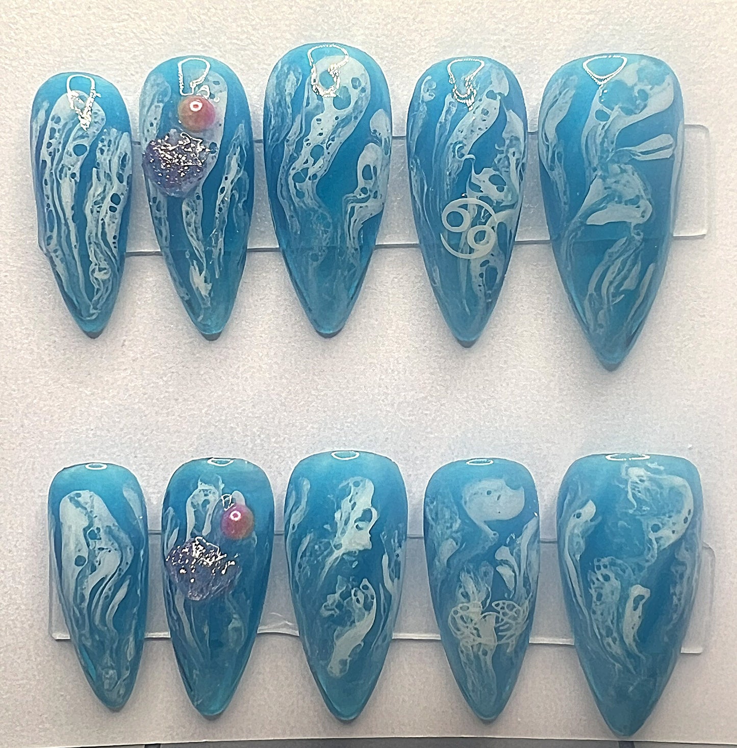 Medium stiletto-shaped press-on nails crafted with a translucent cerulean-blue jelly base and textured white blooming gel waves that look like ocean sea foam. A white Cancer zodiac sign is hidden in the wave pattern on the index finger, and the ring finger is decorated with a 3D purple glitter seashell charm and a round pink-orange pearl accent.