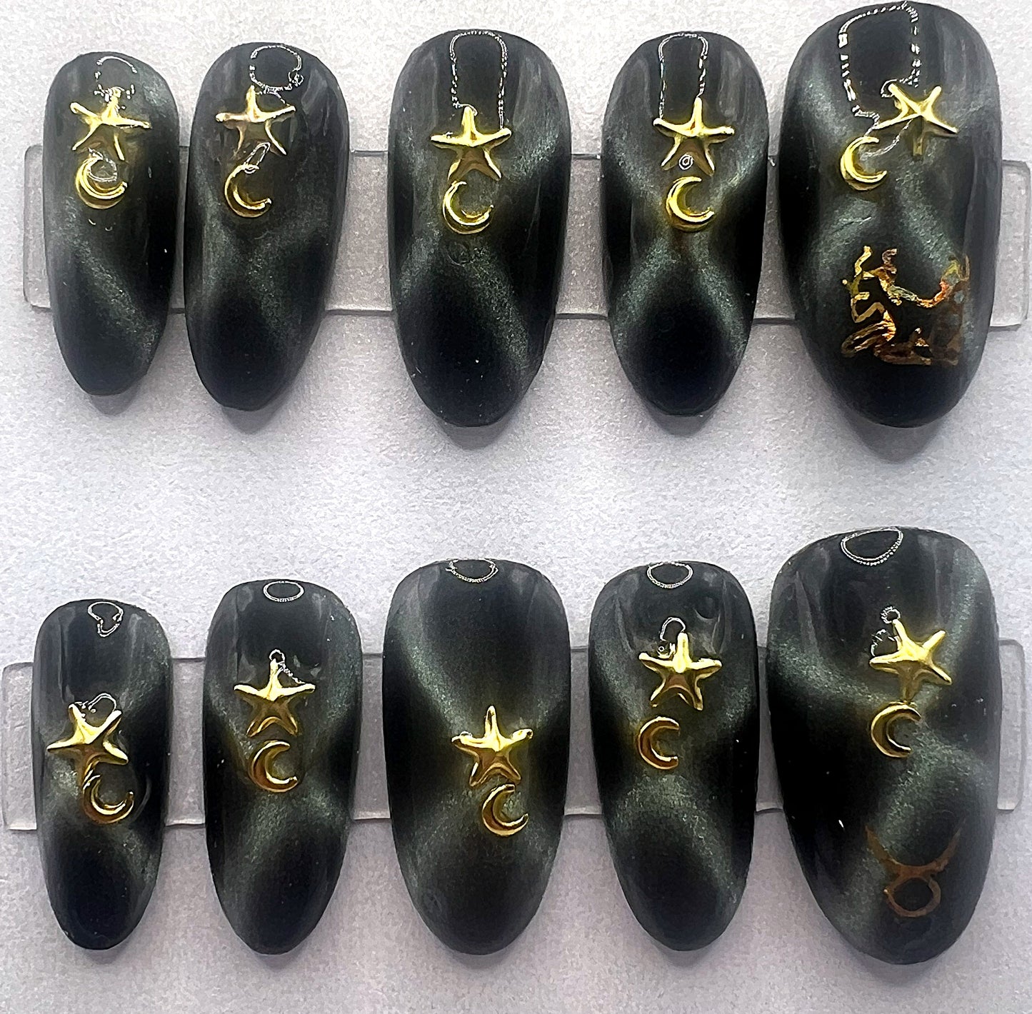 Handcrafted black press on nails with magnetic green gold cat eye effect in an X shape, featuring 3D gold star and moon jewelry charms and a gold holographic Taurus zodiac symbol on the thumb.