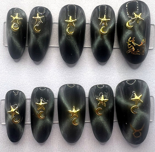 Handcrafted black press on nails with magnetic green gold cat eye effect in an X shape, featuring 3D gold star and moon jewelry charms and a gold holographic Taurus zodiac symbol on the thumb.