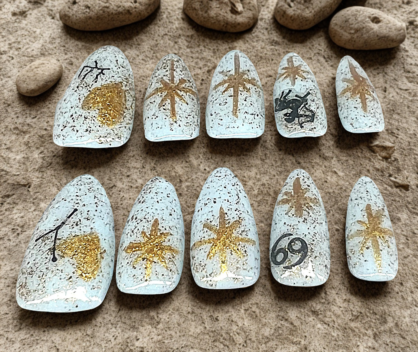 Zodiac Lovers personalized press-on nails featuring a pale blue base with gold foil glitter. The set includes hand-painted gold stars and hearts, with specific black zodiac symbols for Cancer and Sagittarius. Medium almond shape on a stone and sand background.